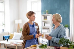 Caregiver chatting with an older woman in a bright kitchen while helping with daily activities