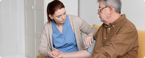 A smiling caregiver in a blue uniform serves a meal on a tray to an elderly woman in a cozy room with large windows and soft lighting.