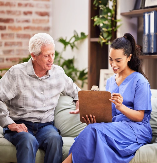 A young nurse in blue scrubs sits on a couch, smiling and holding a clipboard while talking to an older man in a gray shirt.