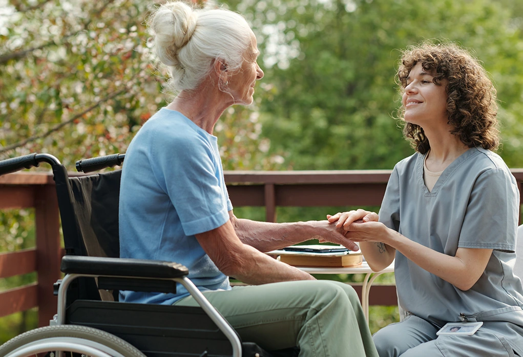 An elderly woman in a wheelchair smiles at a caregiver holding her hand