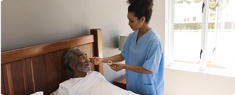 A caregiver in scrubs tends to an elderly man in bed, adjusting his blanket. Soft light from a window creates a calm, caring atmosphere.
