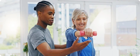 A trainer assists a smiling woman lifting a pink dumbbell indoors. The setting is bright and airy, conveying encouragement and focus on fitness.
