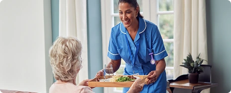 A smiling caregiver in a blue uniform serves a meal on a tray to an elderly woman in a cozy room with large windows and soft lighting.