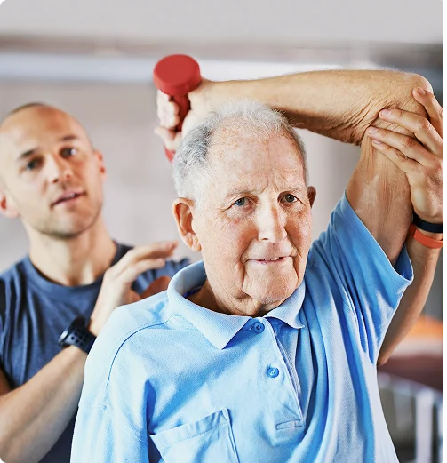 Elderly man in a light blue polo shirt lifts a red dumbbell overhead, assisted by a male trainer. Scene conveys support and focus.
