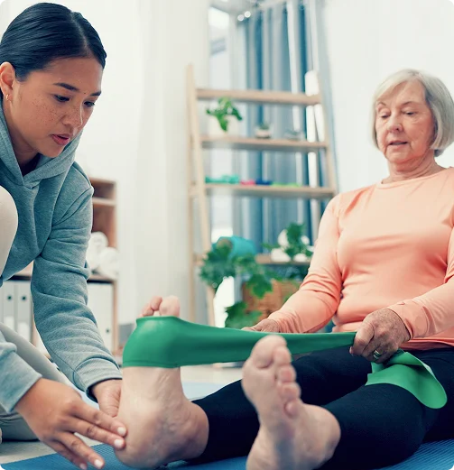 Physical therapist assisting elderly woman with leg stretching exercise using resistance band during rehabilitation session