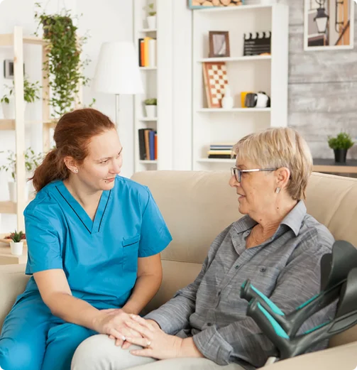 Caregiver in medical scrubs providing emotional support to elderly woman sitting on sofa at home
