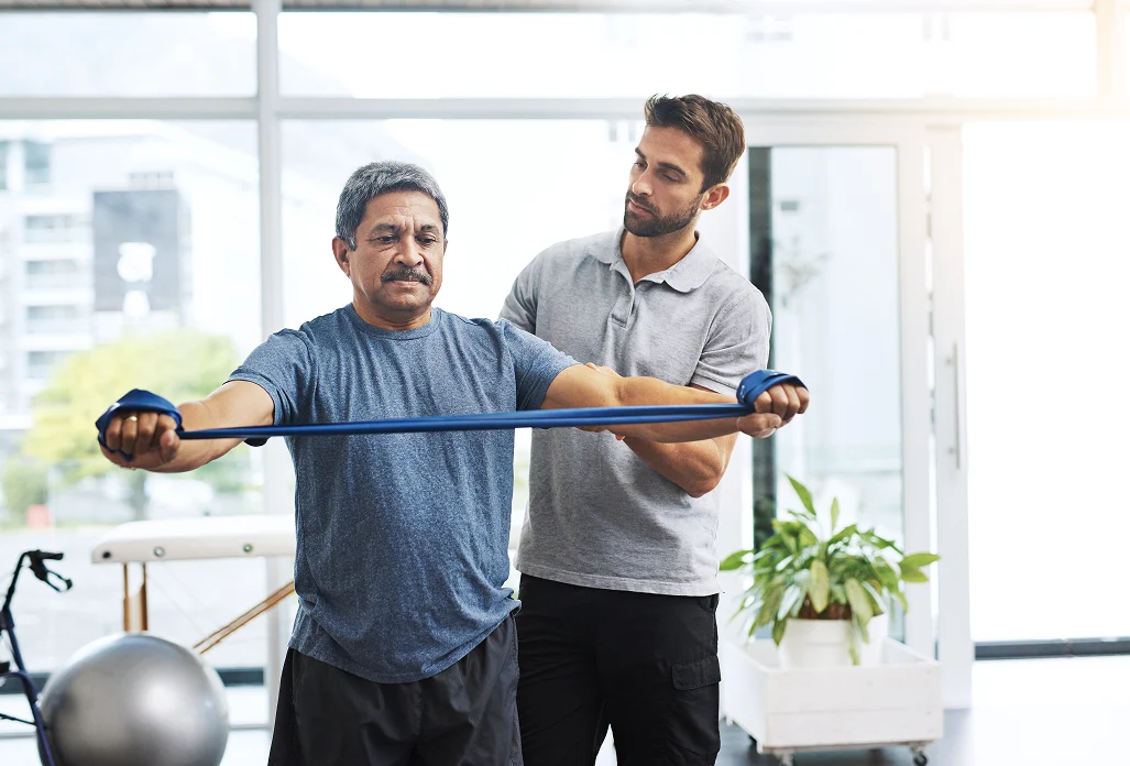 Physical therapist supporting senior man during upper body resistance band exercise in rehabilitation center