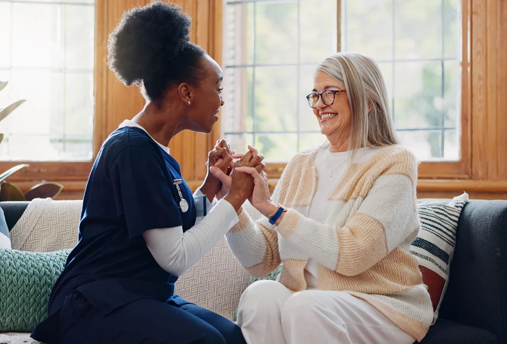 A healthcare worker in navy scrubs joyfully interacts with a smiling elderly woman in a cozy living room. Both hold hands, conveying warmth and companionship.