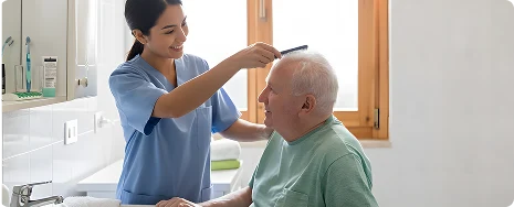 A caregiver in blue scrubs smiles while gently combing the hair of an elderly man in a green shirt, sitting in a bright, sunlit bathroom.
