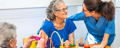 A caregiver in blue scrubs smiles supportively at an elderly woman in a blue top during an engaging group activity at a care facility.