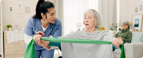 A nurse assists an elderly woman using a green resistance band in a bright room. The woman focuses on the exercise, conveying determination and support.