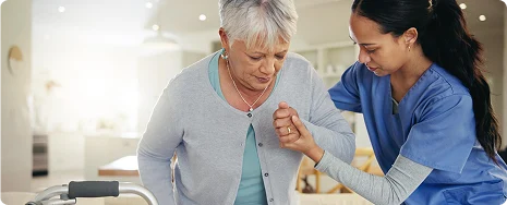 A caregiver in blue scrubs supports an elderly woman with gray hair and a light cardigan as she uses a walker. The setting is a bright room, conveying care and support.