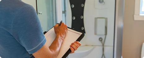 A person in a blue shirt writes on a clipboard while inspecting a modern bathroom with a glass shower. The setting is clean and orderly.
