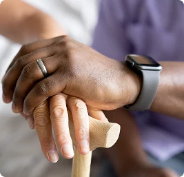 Two hands, one holding a cane, are gently covered by another hand wearing a smartwatch. The image conveys support and care in a comforting setting.