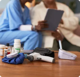 Home healthcare supplies including blood pressure monitor, glucose meter, and medications on table with caregivers in background