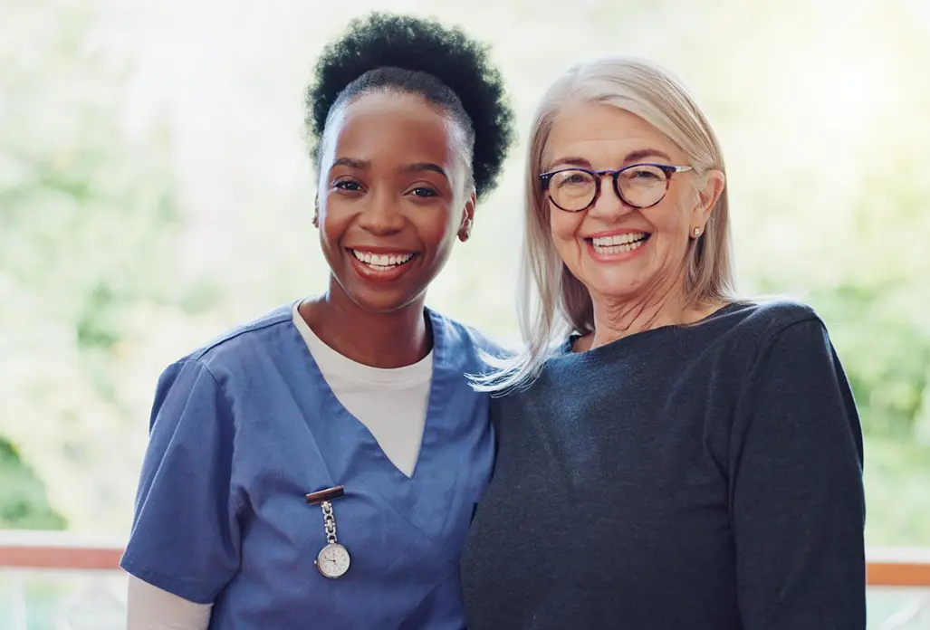 Smiling caregiver standing with an older woman indoors.