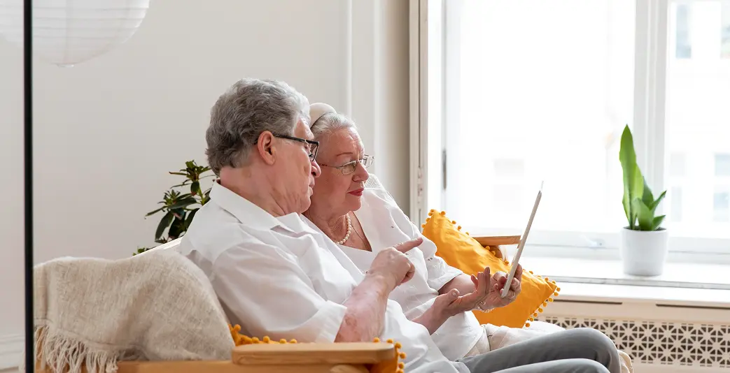 Two elderly people sitting together on a sofa looking at a tablet by a sunny window
