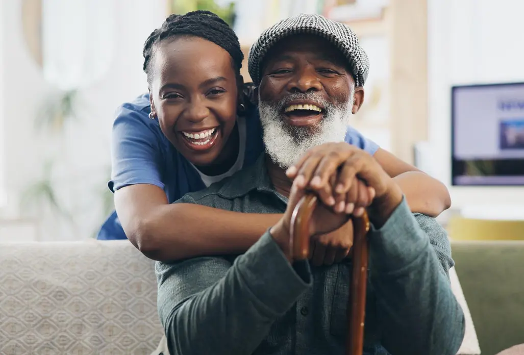 Laughing caregiver in blue scrubs hugging a cheerful elderly man with a cane indoors