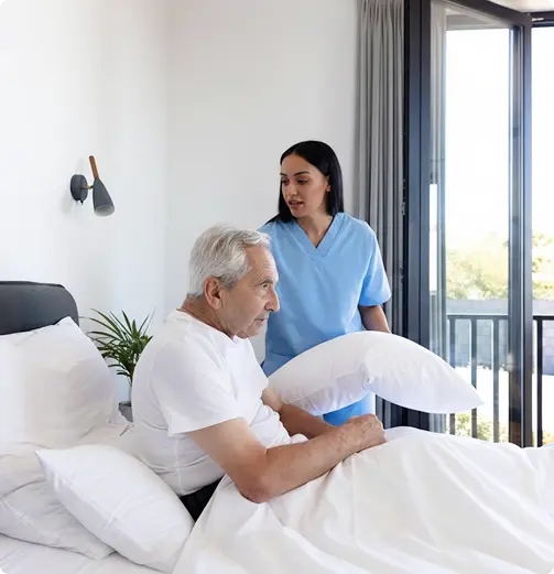 Caregiver in blue scrubs assisting an elderly man sitting up in bed with a pillow