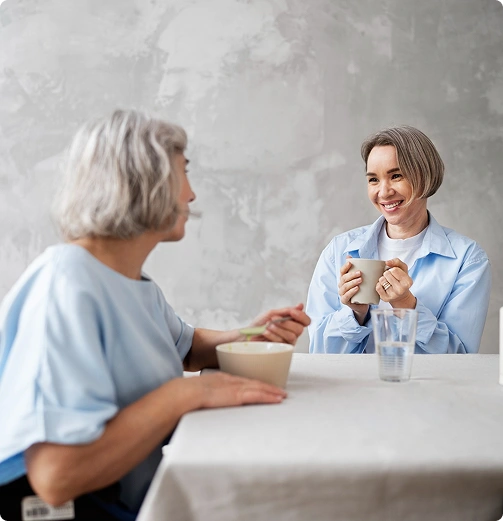 Smiling caregiver sharing a meal and conversation with a senior woman at a table