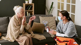 Healthcare professional wearing a face mask discussing medical care with an elderly woman during a home visit.